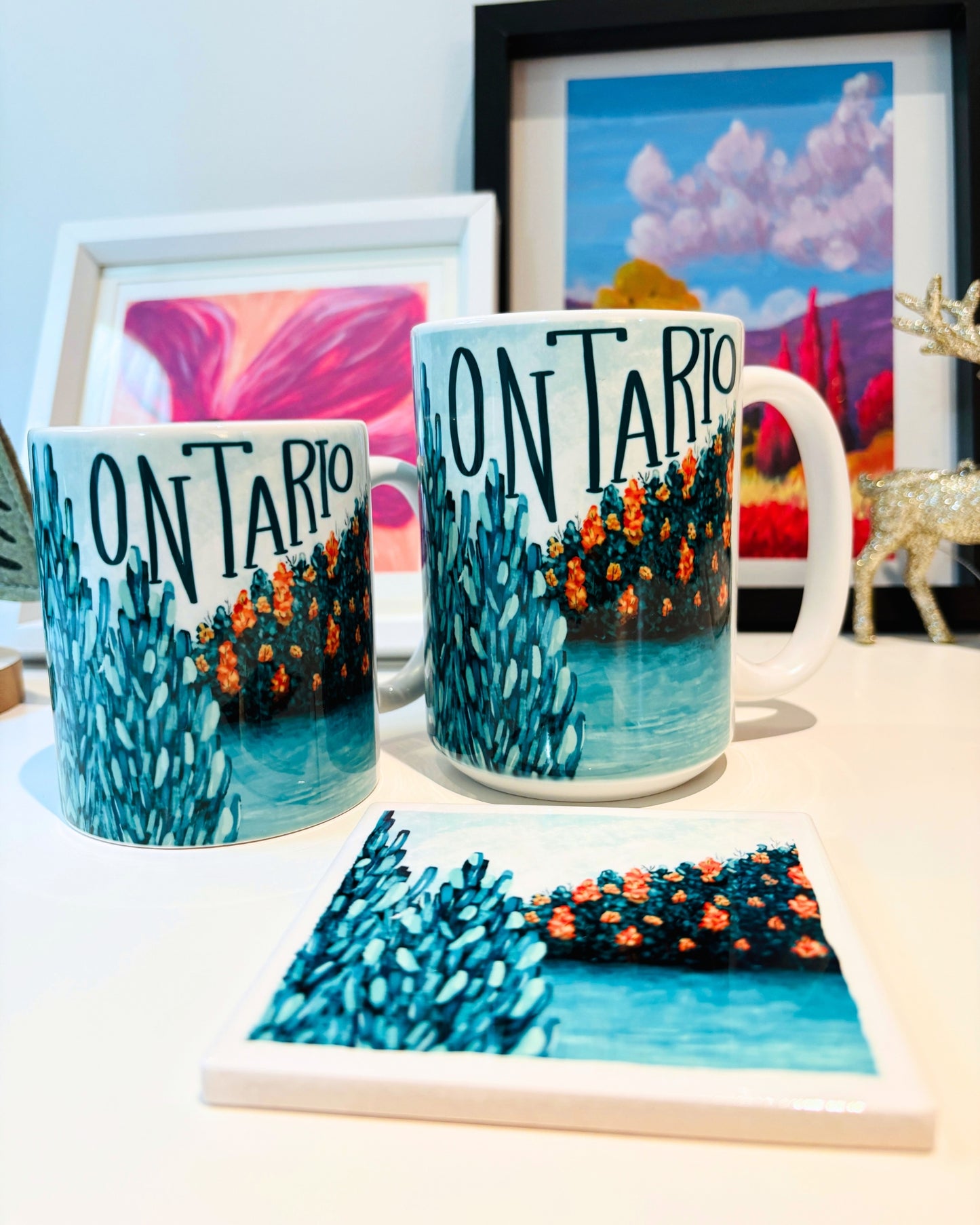 Two mugs and a coaster with 'Ontario' landscape design art on a table with a painting in the background